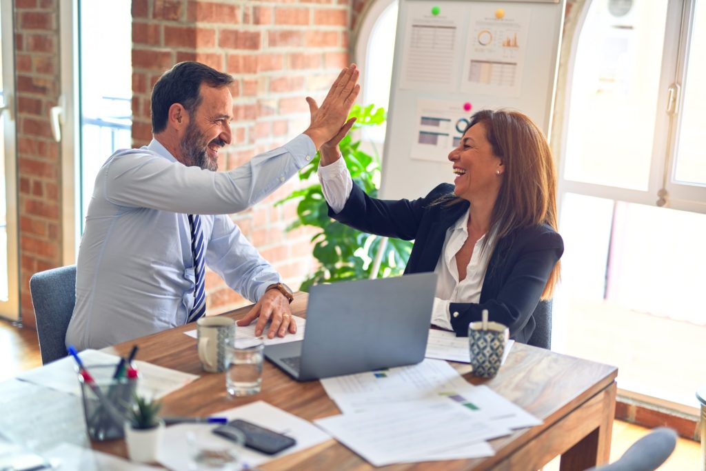 Two marketing colleagues smile and high-five while seated at a meeting table.